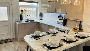 A modern kitchen with a dining area set for a meal, featuring a marble countertop, pendant lights, and a coffee-themed backsplash. - Prestwick - 4 bedroom house, off-street parking (Prestwick)