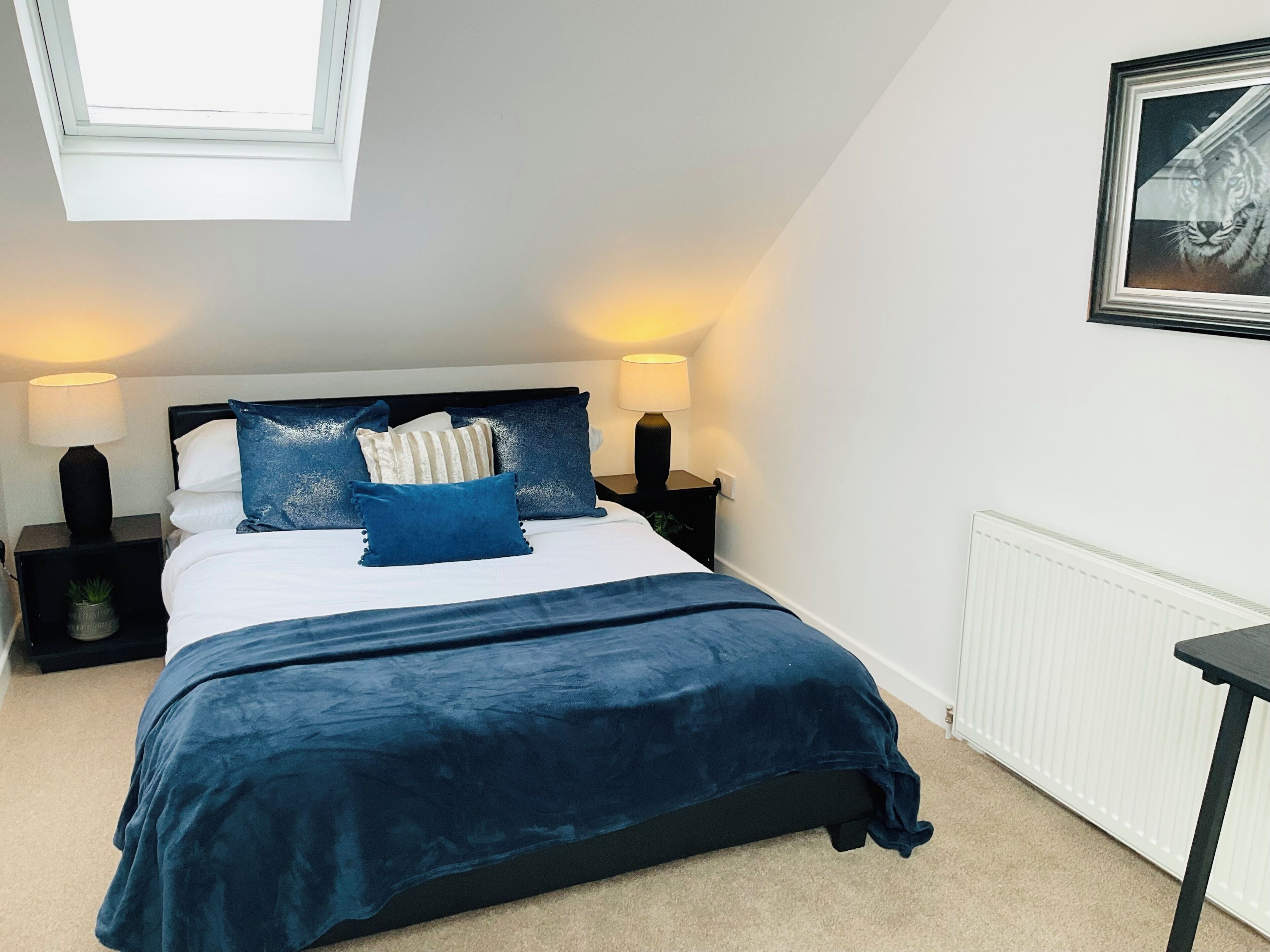 A bedroom with a bed, two bedside tables, a nightstand, a radiator, and a picture on the wall.
