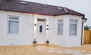 A single-story house with a white exterior, a brown tiled roof, and a front door with two potted plants.