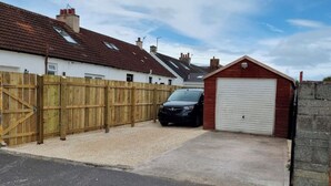 A residential garage with a car parked inside, a wooden fence, and a red garage door. - Prestwick - 4 bedroom house, off-street parking (Prestwick)