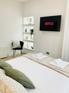 A hotel room with a bed, a chair, a shelf with decorative items, and a TV displaying 'NETFLIX'.