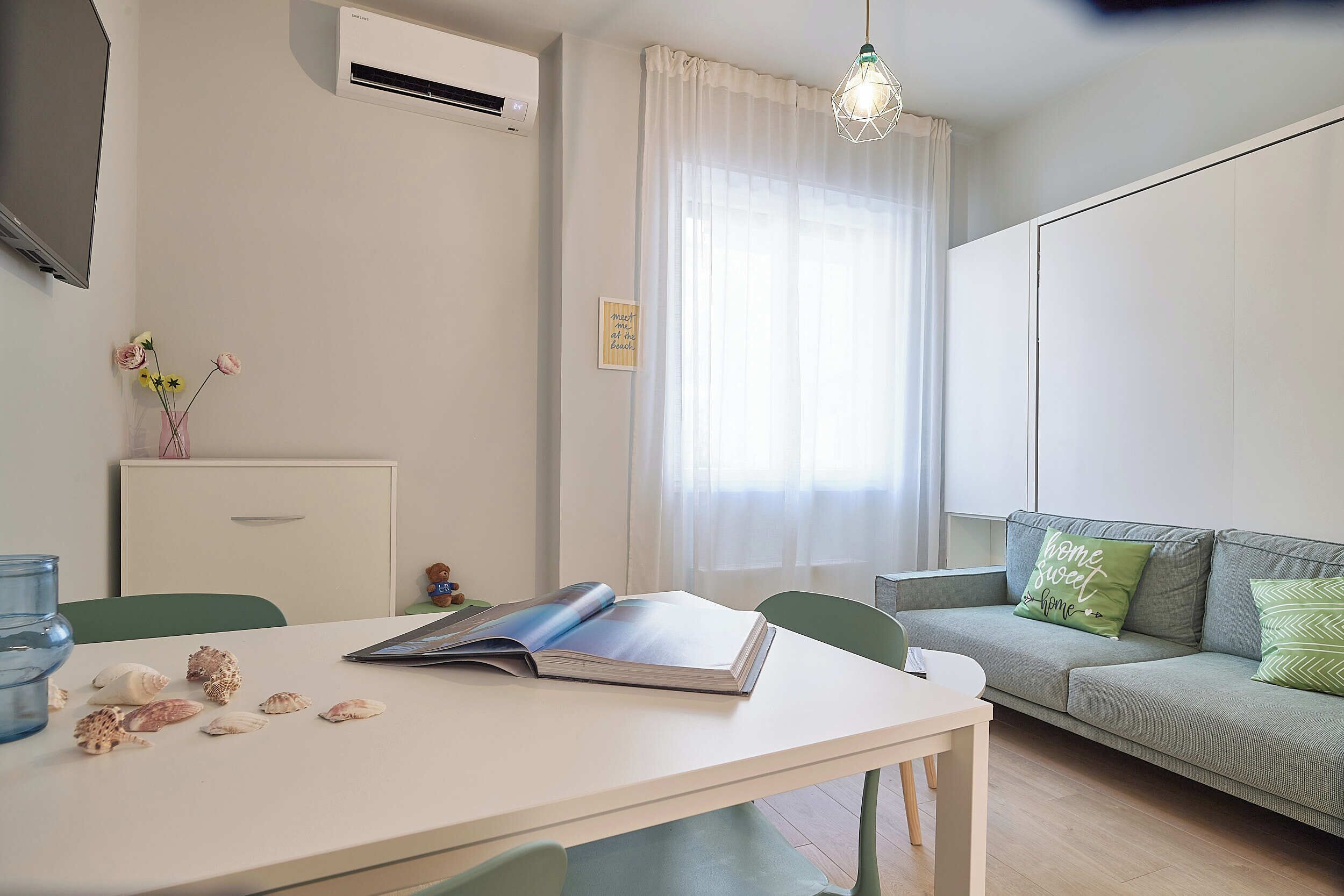 Ein modernes Wohnzimmer mit einem weißen Tisch, einem grauen Sofa mit grünen Kissen, einer Pendelleuchte und einer Wand-Klimaanlage.