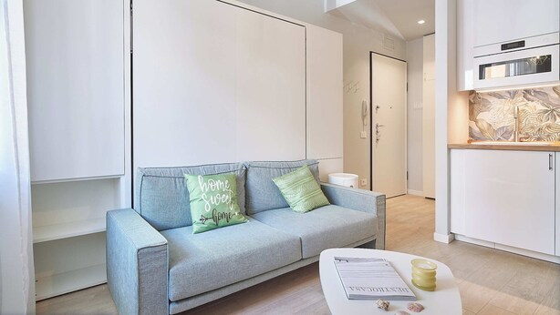 A modern living room with a grey sofa, two pillows, a white coffee table with a book and a candle, and a kitchen area with white cabinets and a microwave. - Mare e Conchiglie (Albenga)