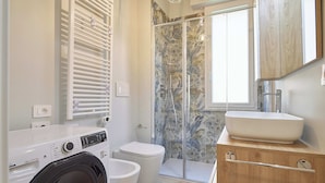 A modern bathroom with a built-in washing machine, a toilet, a shower with a glass door, and a wooden vanity with a sink. - Mare e Conchiglie (Albenga)
