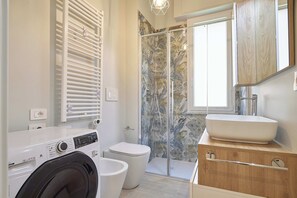 A modern bathroom with a built-in washing machine, a toilet, a shower with a glass door, and a wooden vanity with a sink. - Mare e Conchiglie (Albenga)
