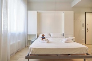 A modern hotel room with a bed, a bedside table, a small bedside lamp, a teddy bear, and a bathroom with a toilet and a shower. - Mare e Conchiglie (Albenga)