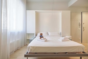 A modern hotel room with a bed, a bedside table, a small bedside lamp, a teddy bear, and a bathroom with a toilet and a shower.