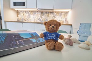 A brown teddy bear with a blue shirt sitting on a table with a framed picture, surrounded by seashells and a blue glass vase.