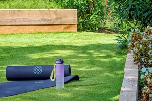 Eine Yogamatte und eine Wasserflasche liegen auf einem gepflegten Rasen mit einem Holz-Pflanzgefäß und blühenden Pflanzen.