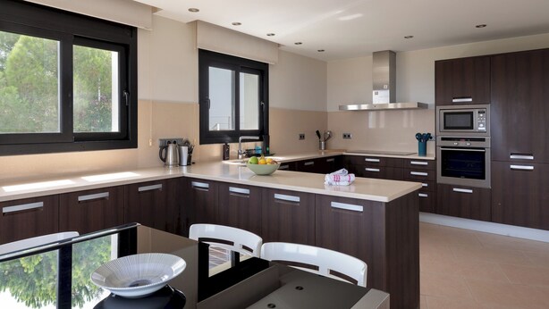 A modern kitchen with dark wood cabinets, a central island with a sink, and built-in appliances including a microwave and oven. - Can Masarol: Views, Pool & BBQ - Platja d'Aro (Castell-Platja d'Aro)