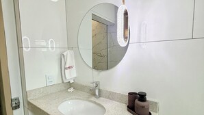 A modern bathroom with a granite countertop, a round mirror, a towel dispenser, and a toilet paper holder.