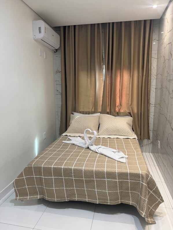 A hotel room with a bed, beige curtains, a white air conditioning unit, and a marble-patterned wall.