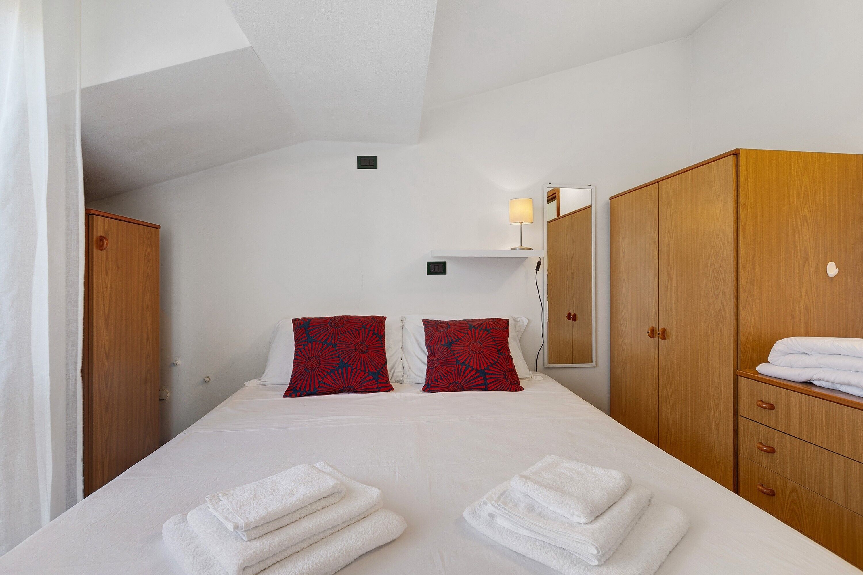 Habitación de hotel con cama, dos almohadas, mesita de noche, lámpara, armario y espejo.