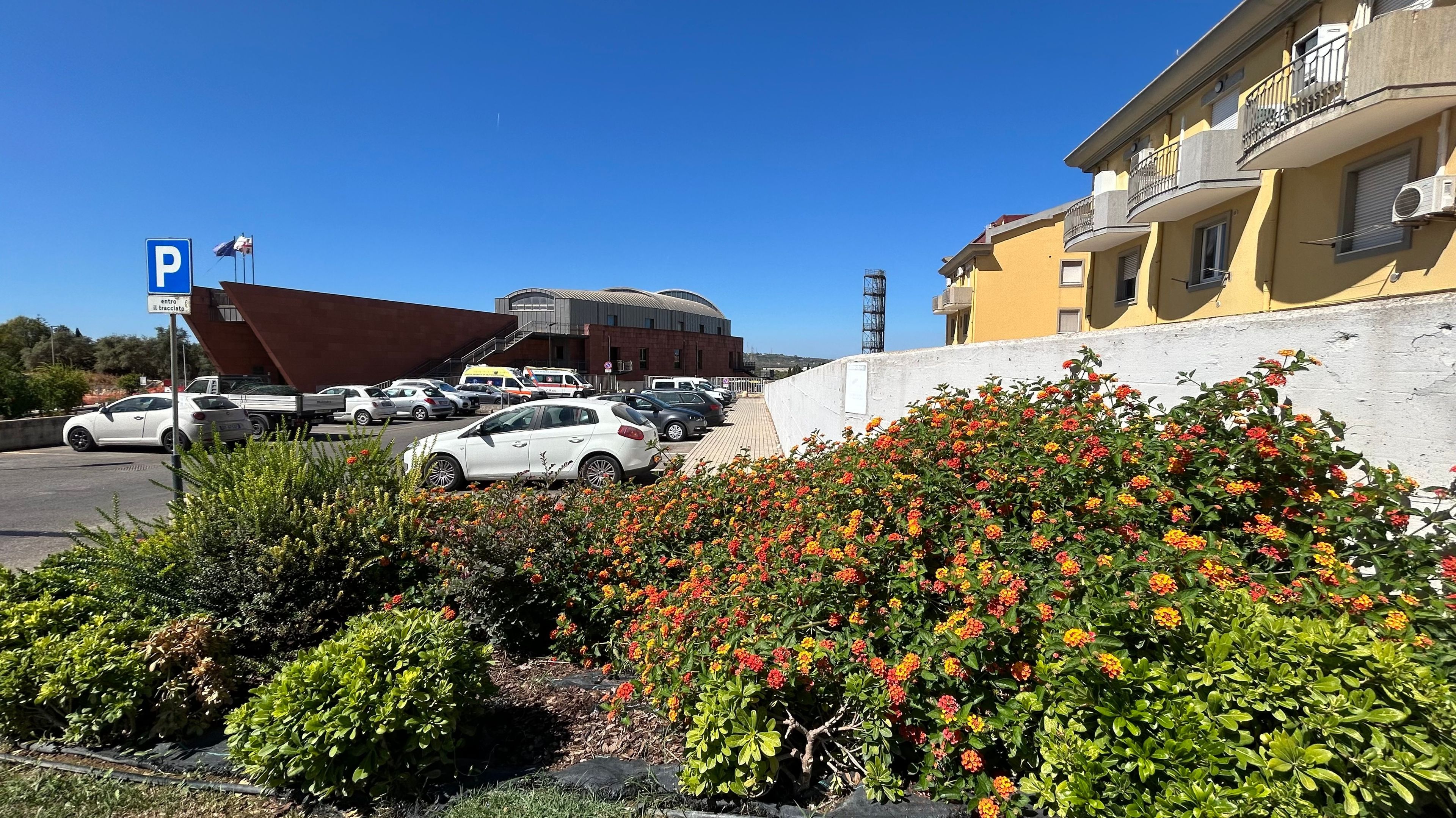 Un estacionamiento con un edificio grande al fondo, cielo despejado y un parterre floral en primer plano.