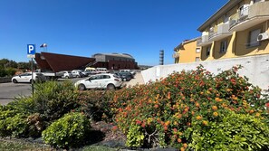 A parking area with a large building in the background, a clear sky, and a flower bed in the foreground.
