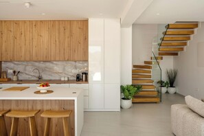 A modern kitchen with wooden cabinets, a marble backsplash, and a central island with stools. - Sirocco Lux Villas (Agios Vasileios)
