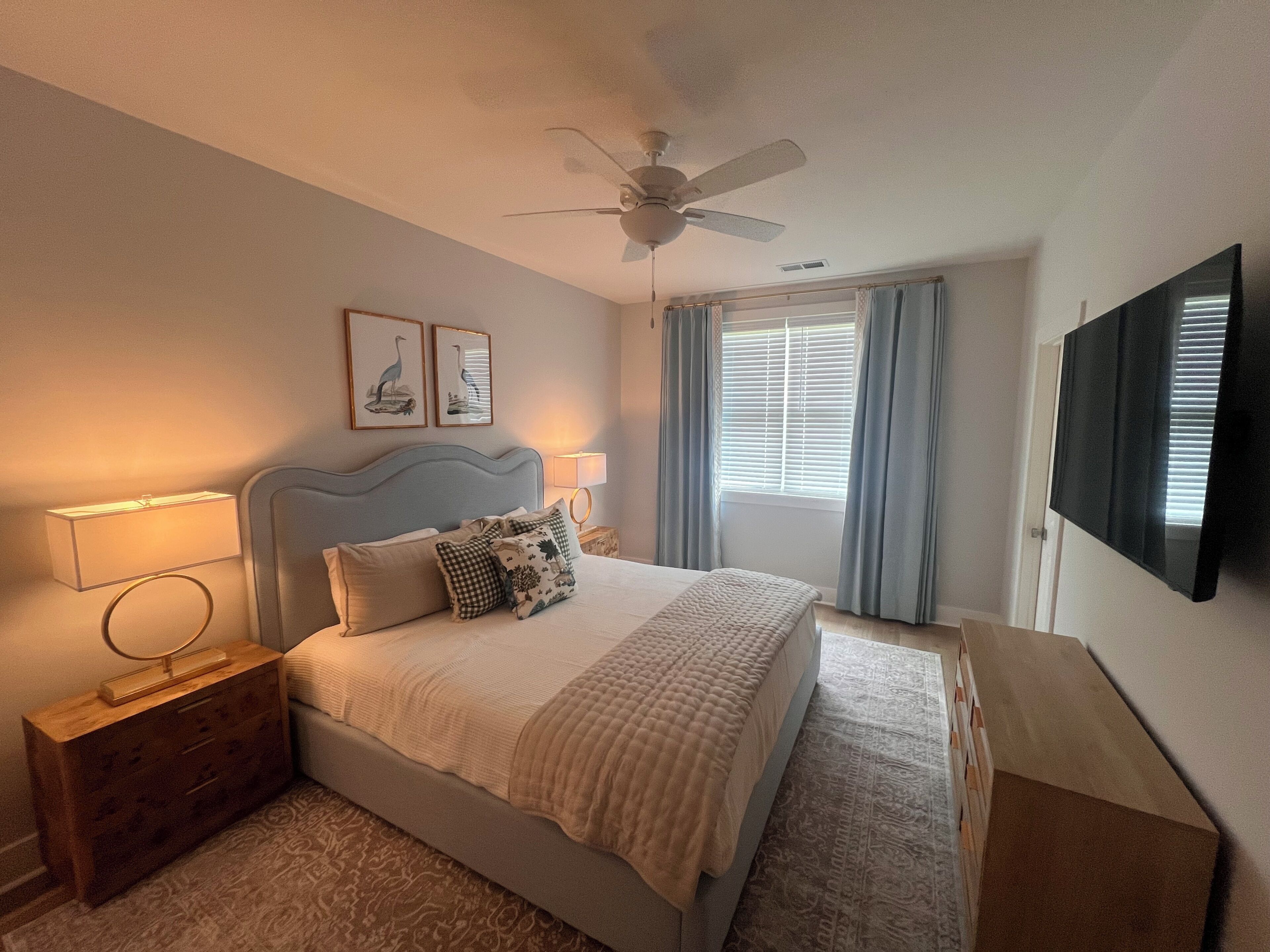 Une chambre à coucher comprenant un lit, des tables de chevet, un ventilateur de plafond, un téléviseur et une fenêtre avec des rideaux.