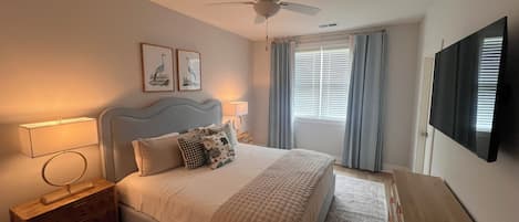 Une chambre à coucher comprenant un lit, des tables de chevet, un ventilateur de plafond, un téléviseur et une fenêtre avec des rideaux.