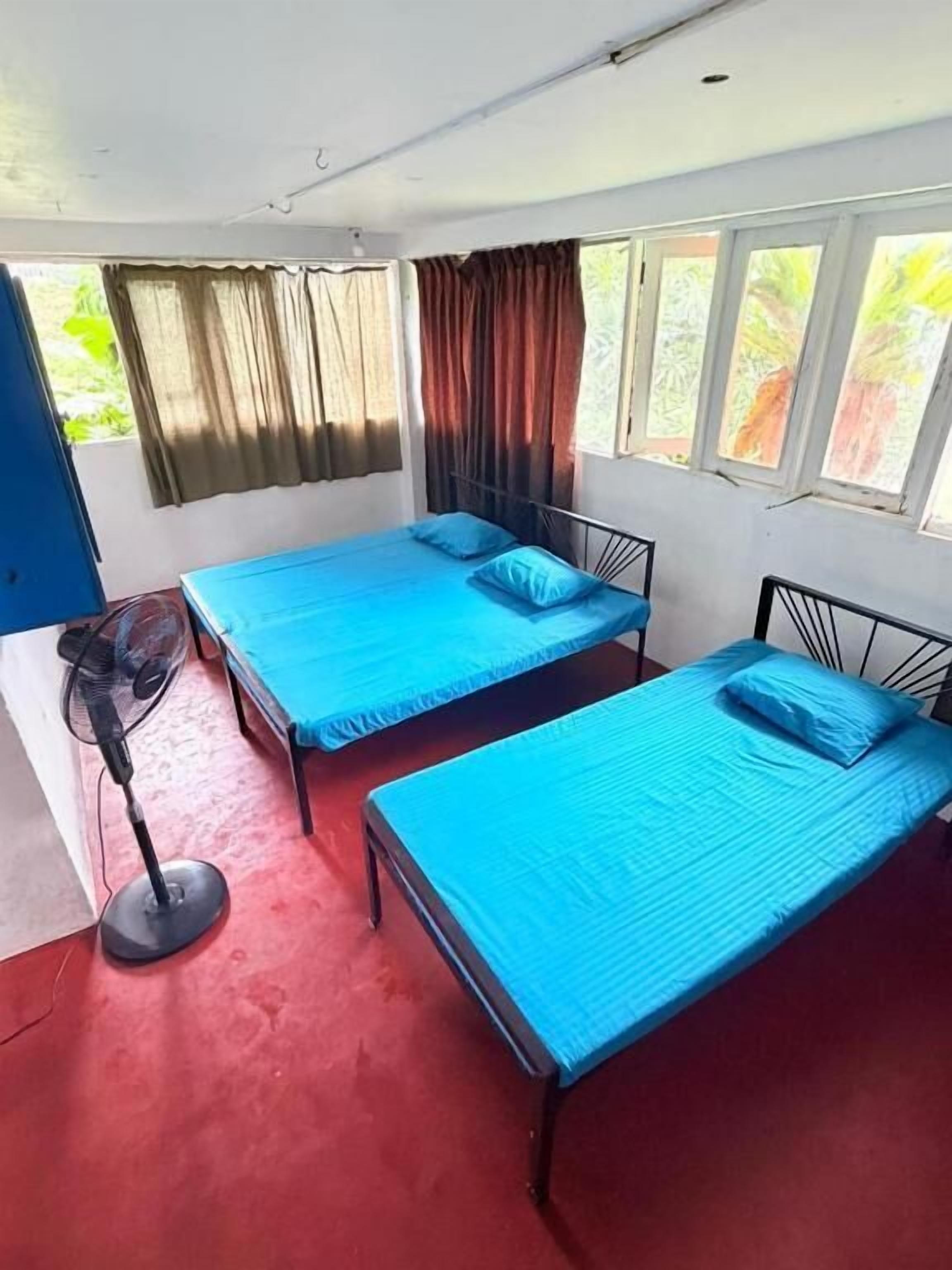Une chambre avec deux lits, un ventilateur sur pied et une fenêtre avec des rideaux.