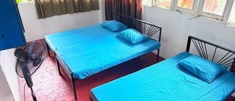 A room with two beds, a standing fan, and a window with curtains.