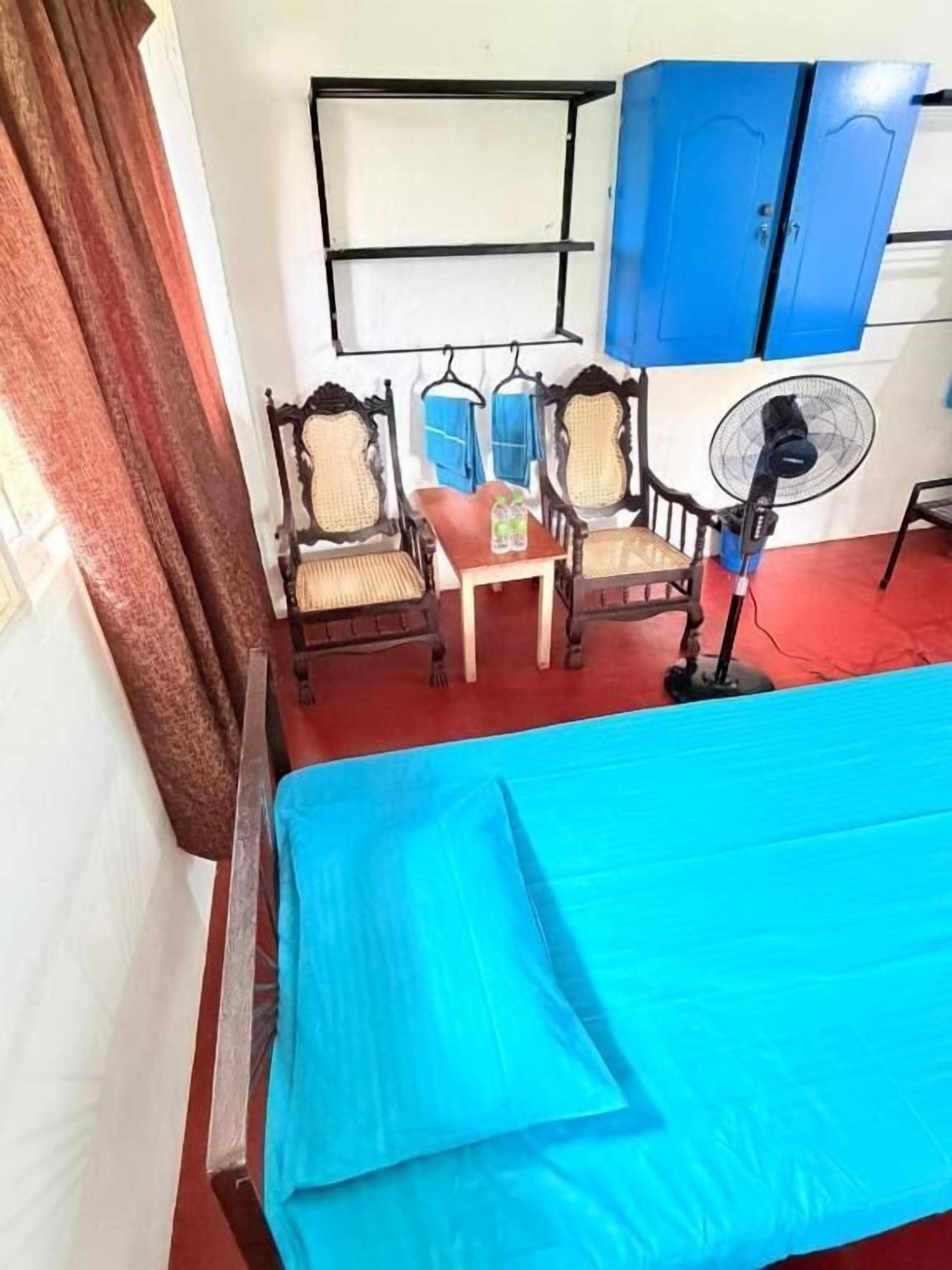 A room with a bed, two chairs, a table, a fan, and a blue cabinet.