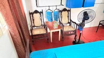 A room with a bed, two chairs, a table, a fan, and a blue cabinet.