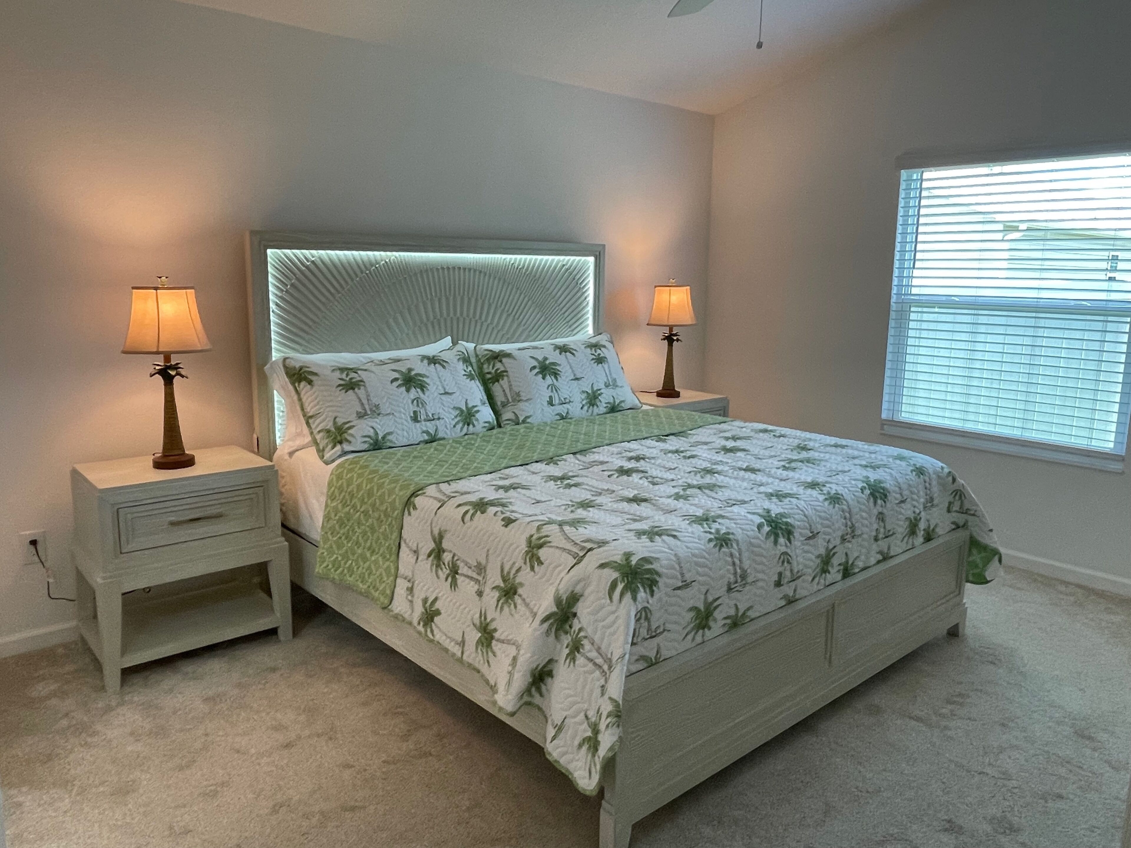 A bedroom with a bed, two bedside tables, lamps, a window with blinds, and a carpeted floor.