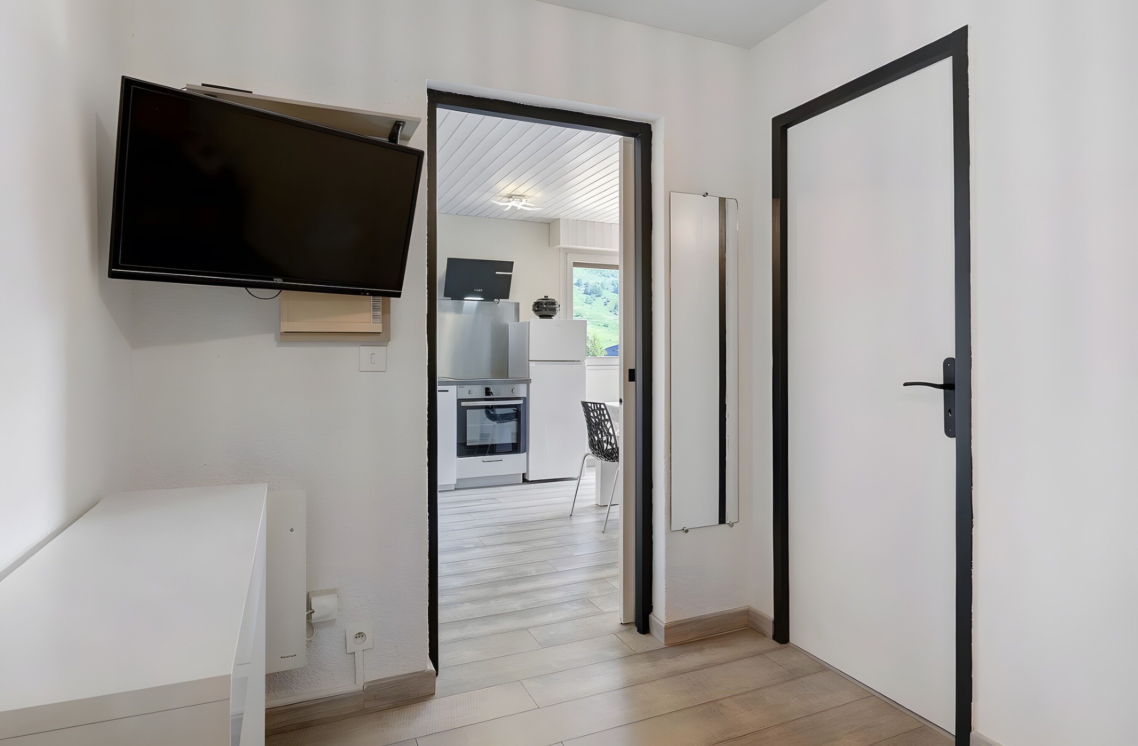 Un couloir avec un téléviseur fixé au mur, une porte donnant sur une cuisine et une vue sur l’extérieur.