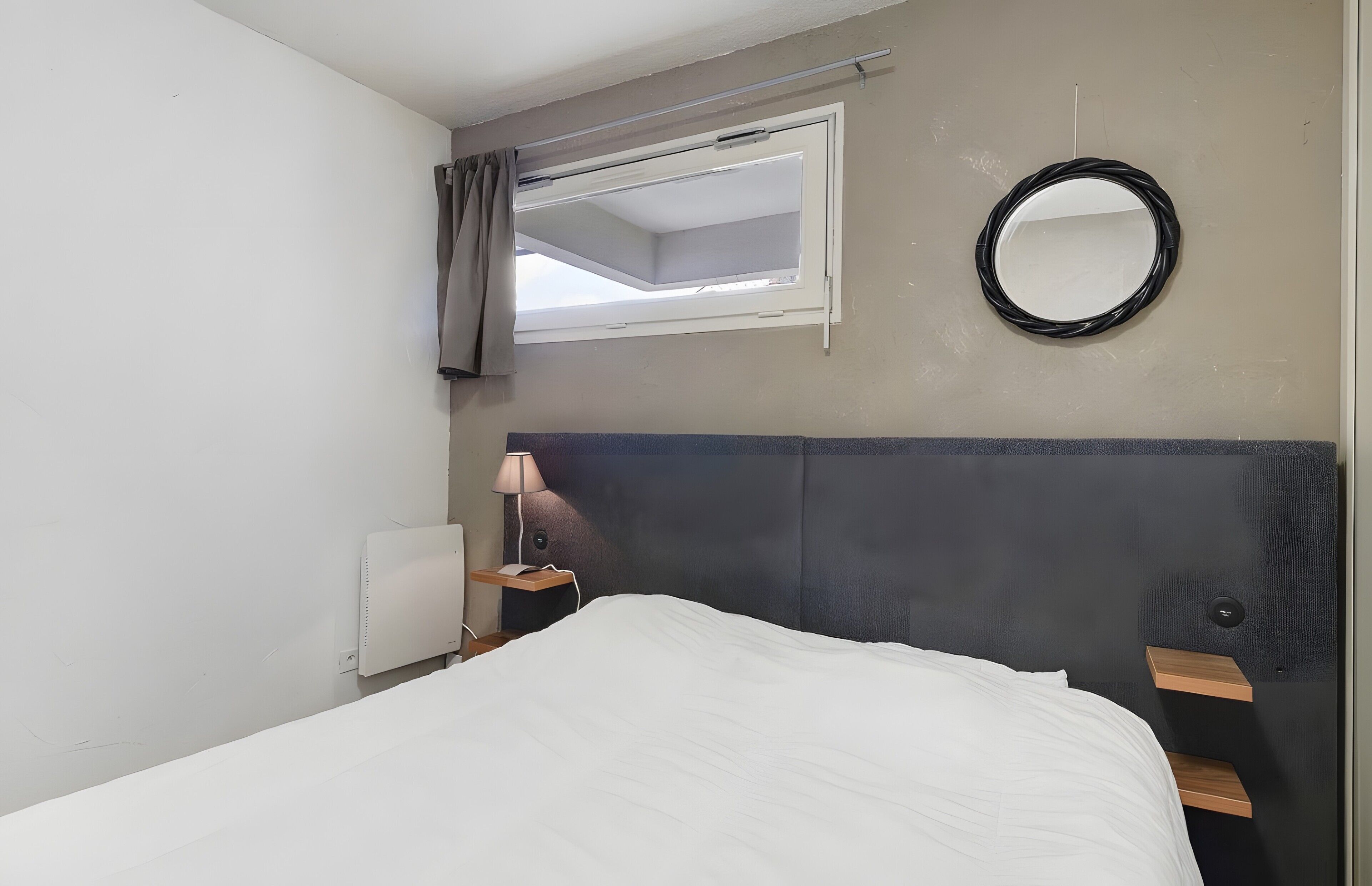 Une chambre d’hôtel comprenant un lit, une table de chevet, une lampe, une fenêtre avec des rideaux et un miroir fixé au mur.