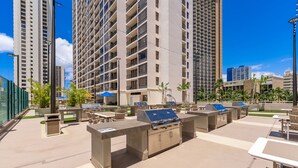 An outdoor patio area with barbecue grills, tables, and chairs, surrounded by tall buildings. - Waikiki Banyan, Top Floor Oceanview Pool + Parking (Honolulu)