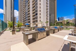 An outdoor patio area with barbecue grills, tables, and chairs, surrounded by tall buildings. - Waikiki Banyan, Top Floor Oceanview Pool + Parking (Honolulu)
