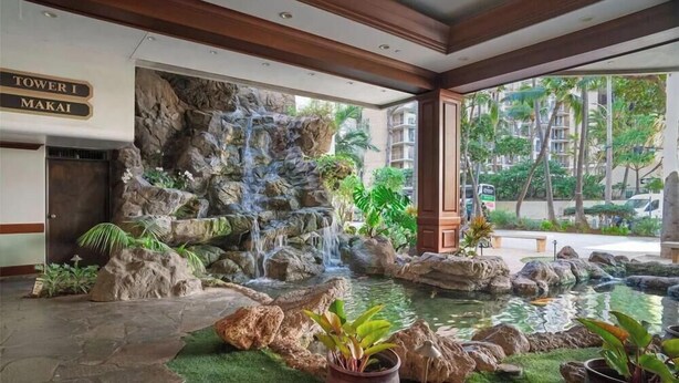 A hotel lobby with a waterfall feature, a sign for 'Tower I Makai', and a view of the surrounding buildings. - Waikiki Banyan, Top Floor Oceanview Pool + Parking (Honolulu)