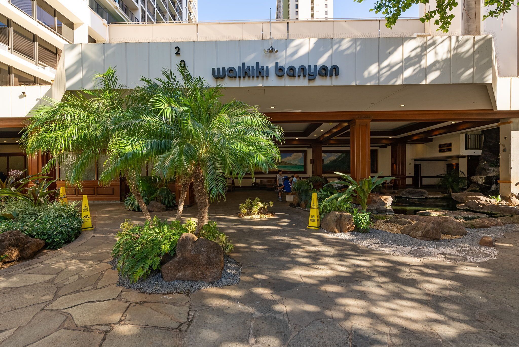 L’entrée d’un hôtel, avec une enseigne indiquant « Wakiki Banyan », et un espace paysager agrémenté de palmiers et de rochers.