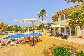 A poolside area with lounge chairs and umbrellas, a pool, and a building with a balcony. - Villa Panoramick - Plusholidays (Calpe / Calp)