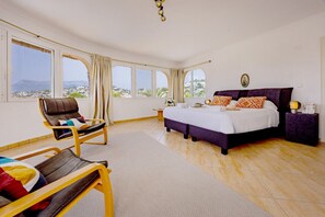 A bedroom with a bed, a chair, a bedside table, and large windows. - Villa Panoramick - Plusholidays (Calpe / Calp)