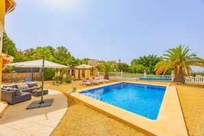 Une piscine entourée de chaises longues et d’un canapé, le tout bordé de palmiers et délimité par une clôture blanche.