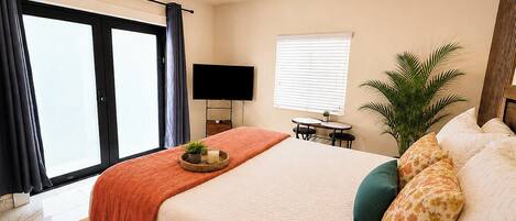 A hotel room with a bed, a television, a small table, a potted plant, and a window with blinds.