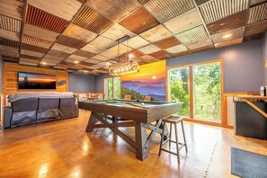 A pool table in a room with a wooden ceiling, a sofa, and large windows with a view of greenery. - Smoky Mtn Home with Theater, Game Room & Hot Tub (Sevierville)