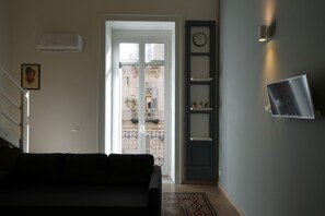 Ein Wohnzimmer mit Sofa, einer Treppe, einer zur Außenseite führenden Tür, einer Wanduhr und einem an der Wand befestigten Fernseher.
