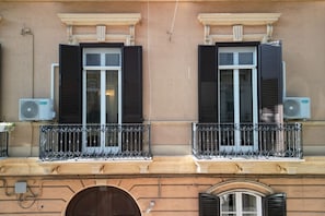 Eine Gebäudefassade mit zwei Fenstern, jedes mit Fensterläden, und einem Balkon mit schmiedeeisernem Geländer.