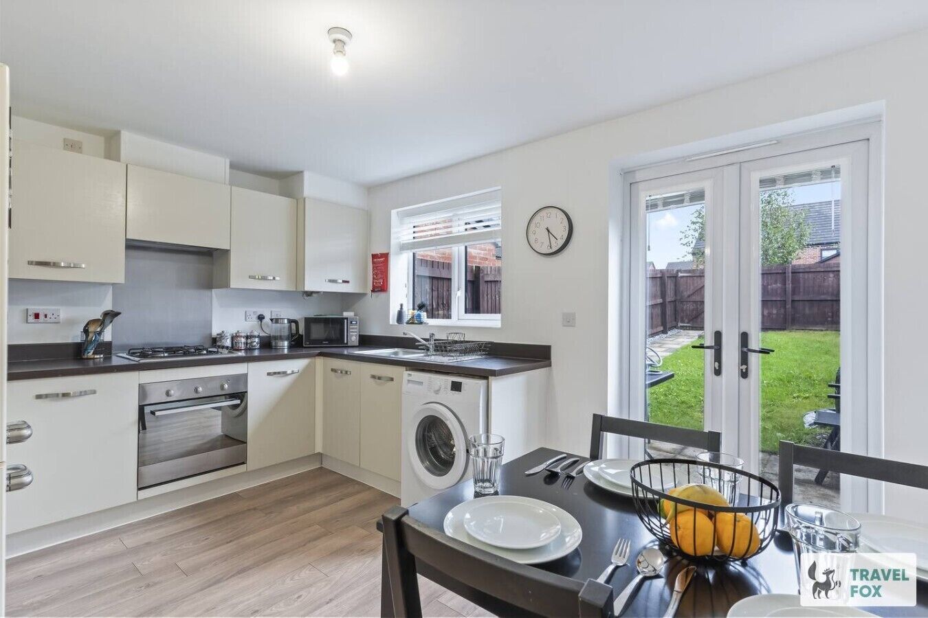 A compact kitchen with a washing machine, a dining area with a table set for two, and a view of a backyard through a sliding door.