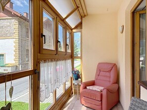 Ein Zimmer mit einem raumhohen Holzfenster, einem rosa Stuhl und Blick auf eine Straße und Berge.
