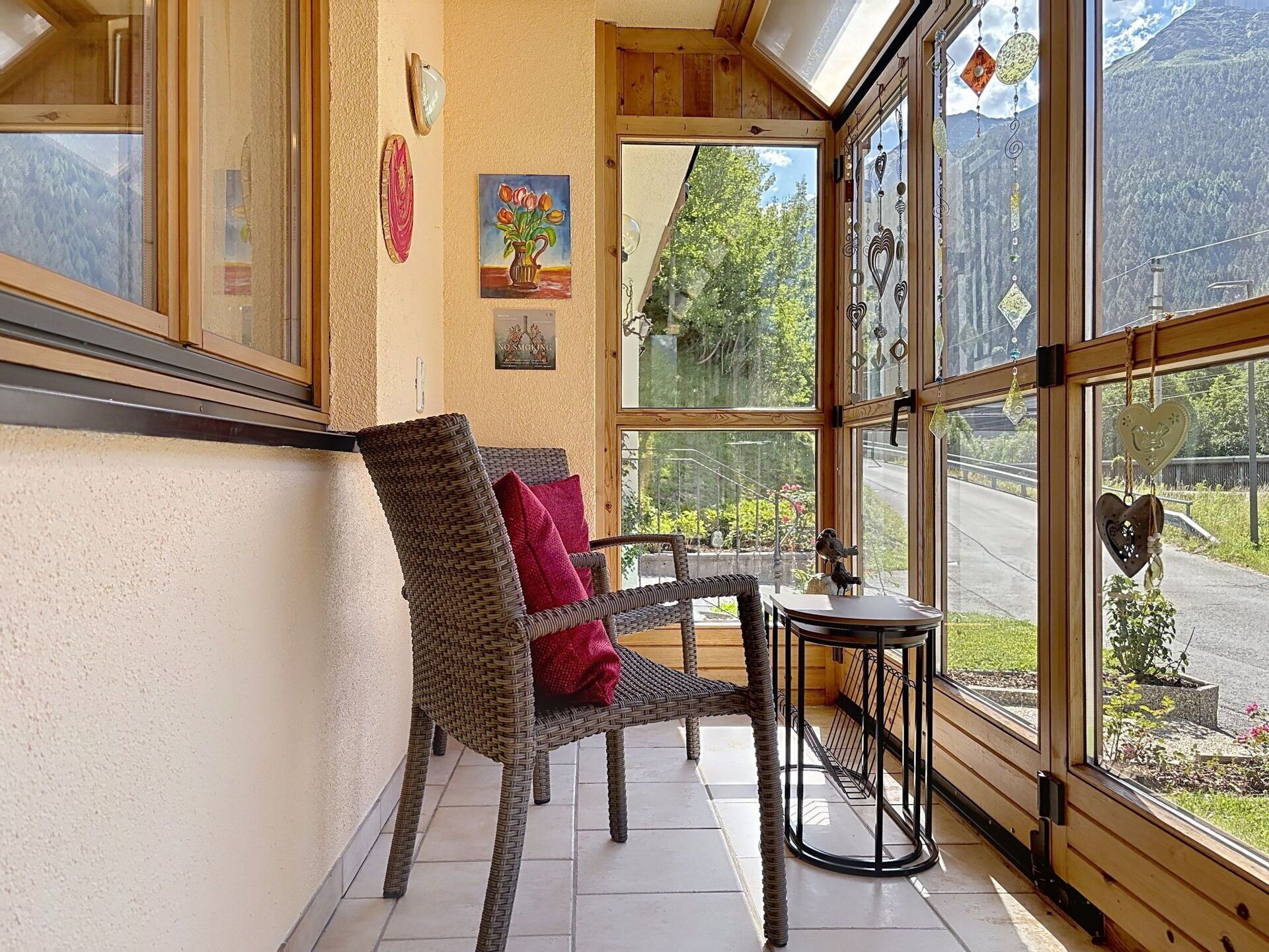 Une pièce accueillante avec un fauteuil en osier, une petite table et une vue sur une route et la verdure, le tout visible à travers de grandes fenêtres.