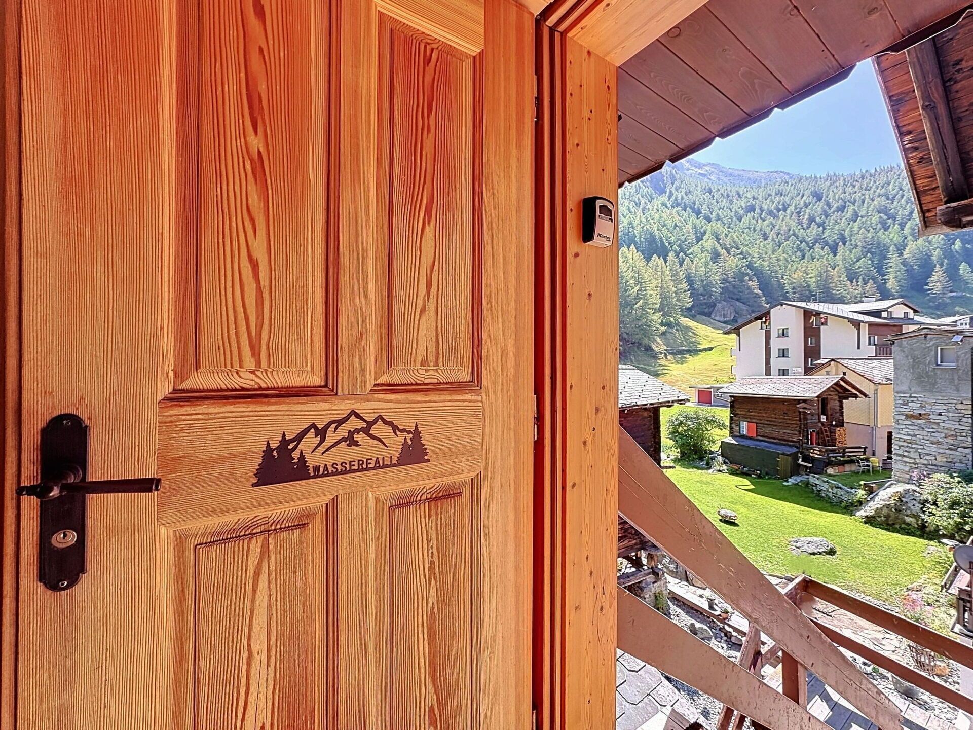 Une porte en bois ornée d’une sculpture représentant un paysage de montagne, une vue sur une région montagneuse avec des bâtiments, et une balustrade en bois.