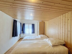 Ein Zimmer mit Holzwandverkleidung, einem Bett mit weißer Bettwäsche, einem Stuhl und einem Fenster mit Vorhängen.