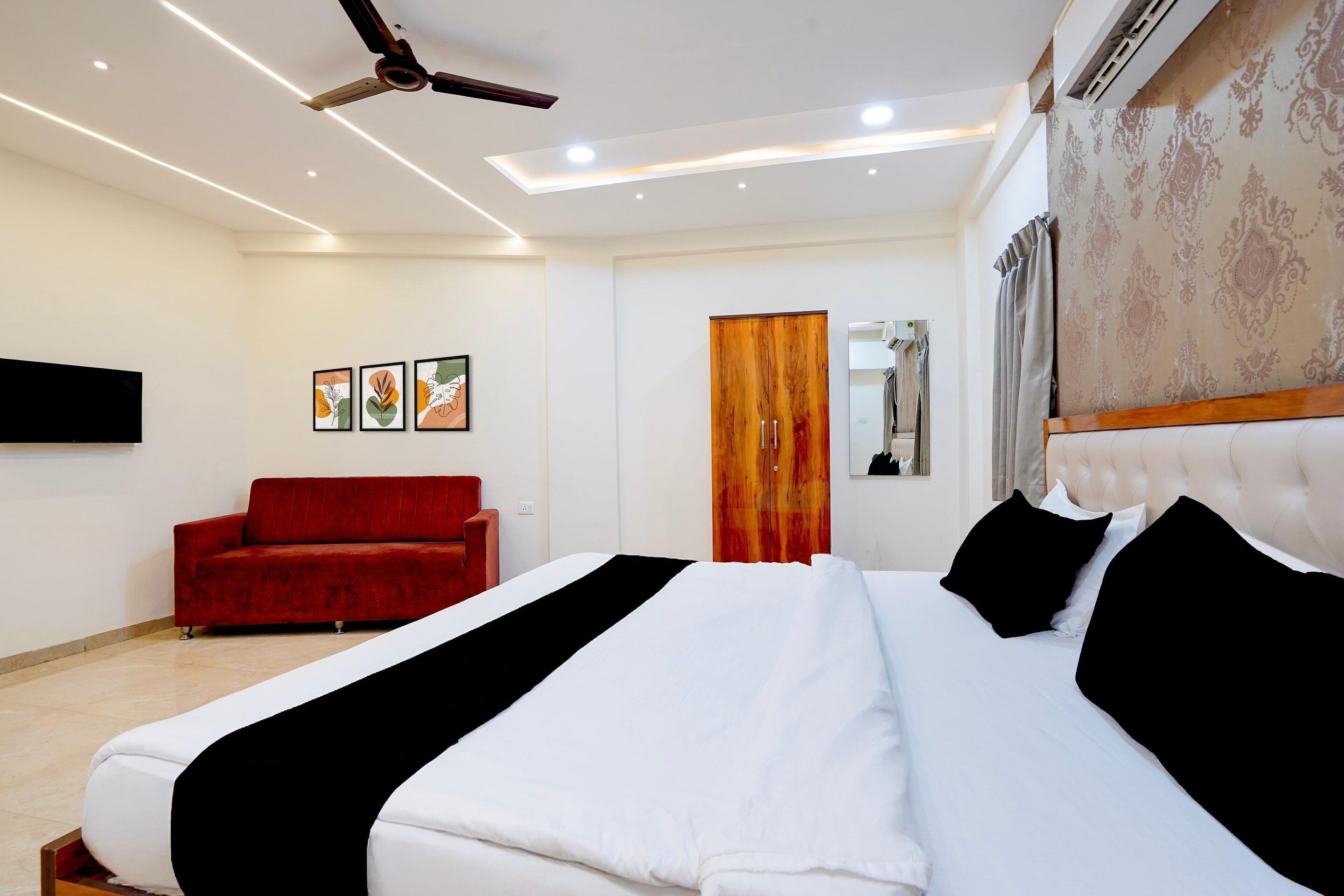 A hotel room with a bed, a red sofa, a ceiling fan, and a wall-mounted TV.
