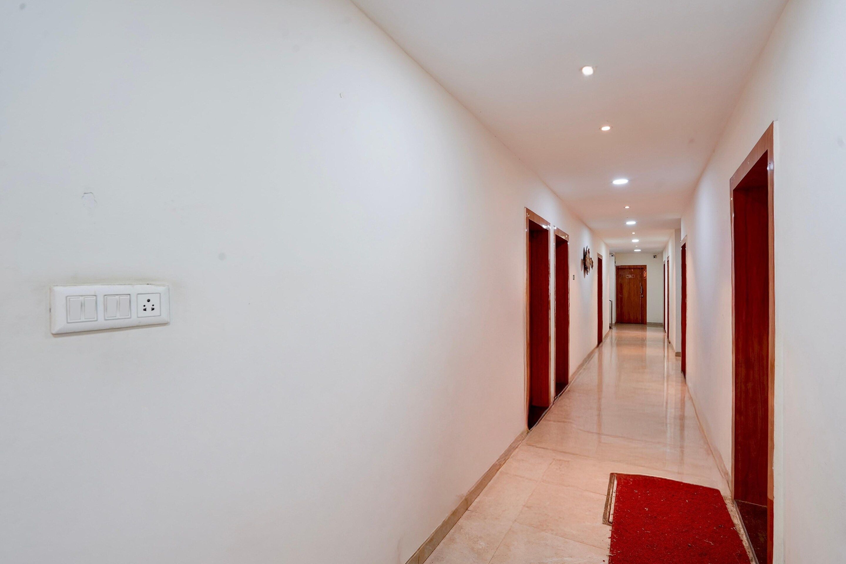 A long, narrow hallway with white walls, a red carpet, and a light switch on the left.