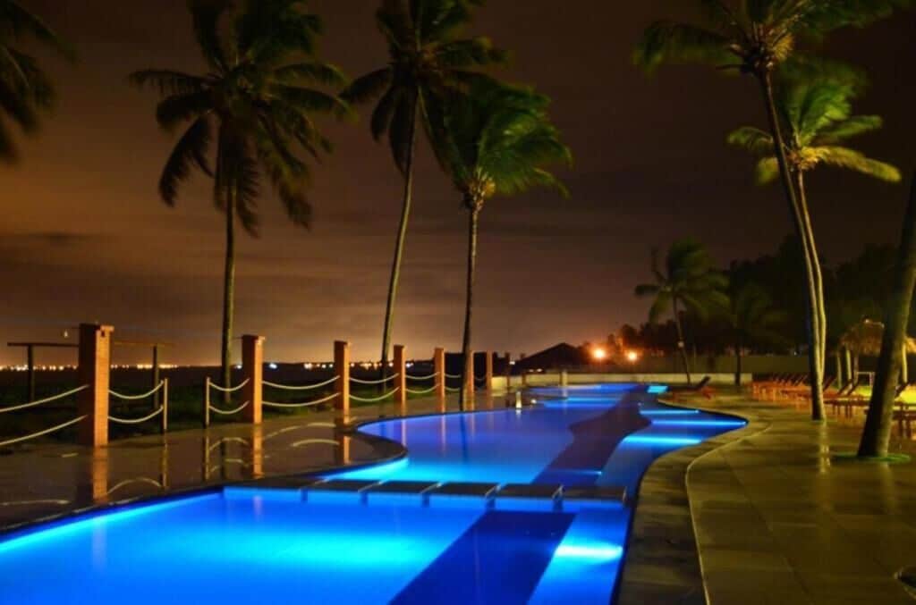 Una piscina iluminada con luces azules, rodeada de palmeras y una valla.