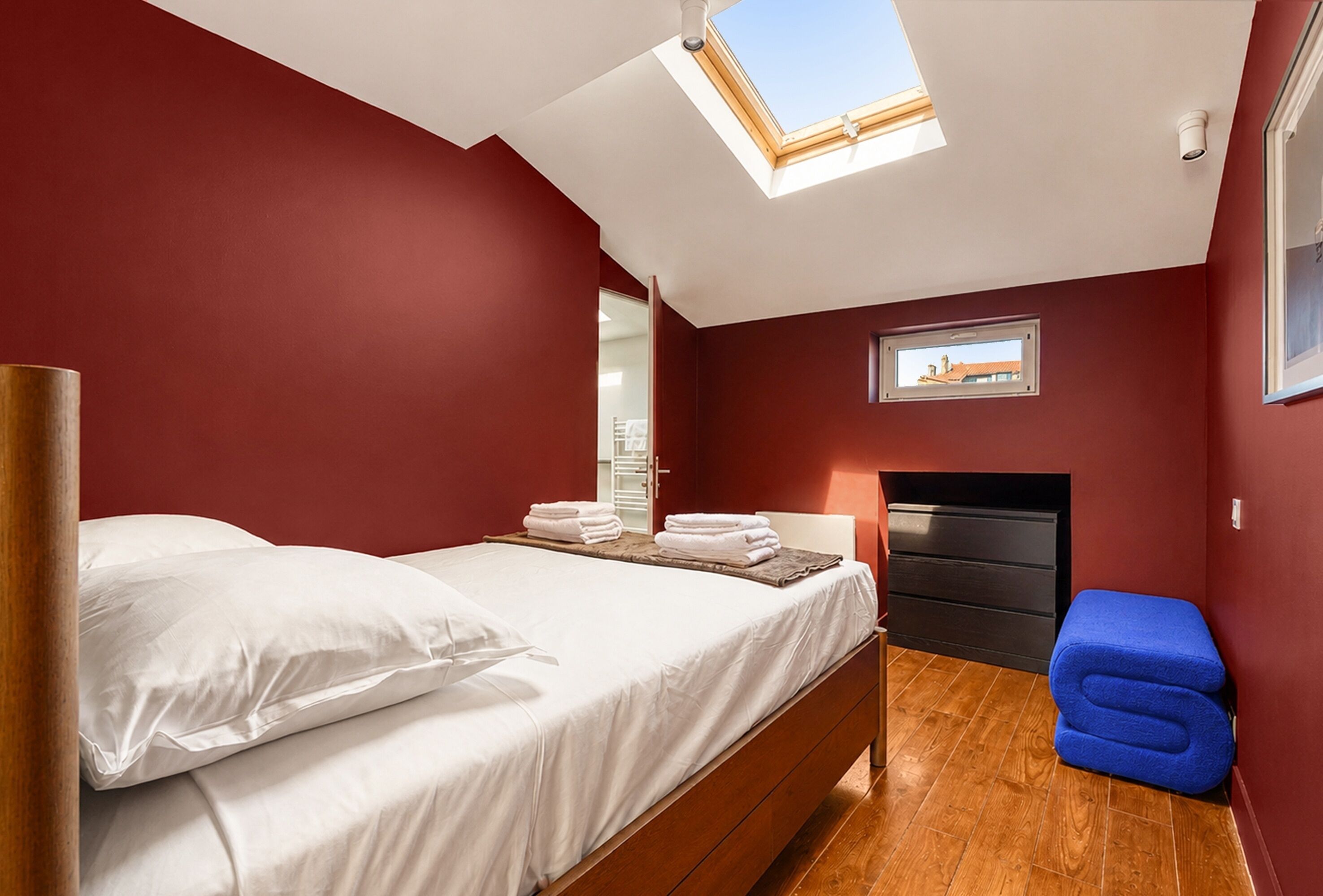 A bedroom with a skylight, a bed with white linens, a blue bench, and a fireplace.