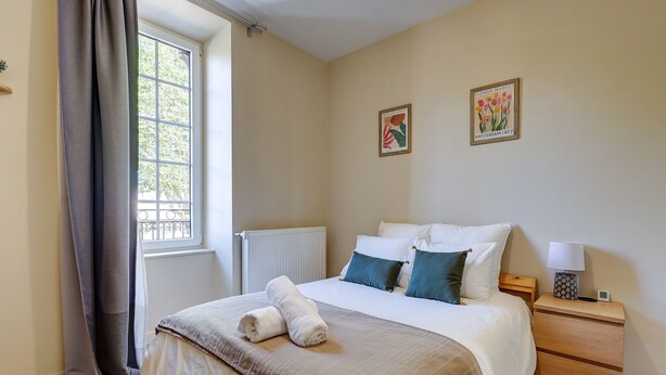 A bedroom with a bed, two pillows, a bedside table with a lamp, a clock, and a window with curtains. - La Maison des Prés - Maison bourgeoise de 1890 (Montceau-les-Mines)